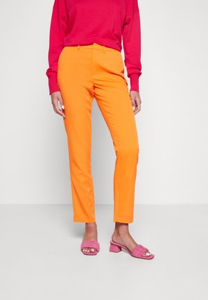 Mujer con pantalones naranja brillantes, una blusa roja de manga larga y sandalias de tacón bloque con punta abierta rosas, de pie sobre un suelo blanco.