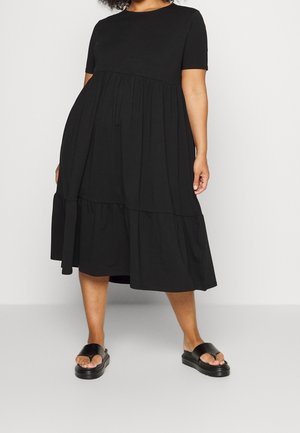Frau trägt ein schwarzes Midi-Kleid mit kurzen Ärmeln und geraffter Taille sowie schwarze Plateau-Sandalen und steht auf einem schlichten hellen Boden.