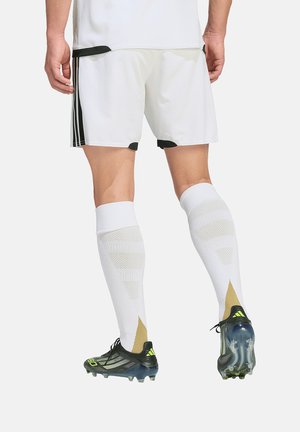 Mężczyzna w białych szortach piłkarskich, długich białych skarpetach i czarnych korkach z zielonymi akcentami, stojący z nogami lekko rozstawionymi, widok od tyłu.