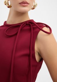 Top bordeaux con una finitura testurizzata, caratterizzato da un dettaglio annodato sulla spalla e un design senza maniche. Orecchini a cerchio dorati audaci accentuano il look.