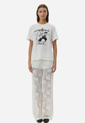 Hvid grafisk t-shirt med sort print, parret med løse, semi-gennemsigtige hvide blondebukser med blomstermønster. Enkle sorte sko.