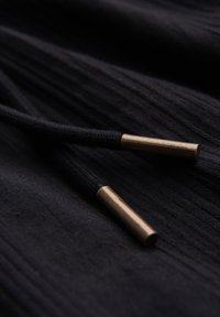 Tissu noir avec plis texturés et deux cordons noirs terminés par des embouts métalliques couleur bronze.