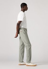Pantaloni di cotone verde chiaro con gamba dritta, tasche laterali e una patch con logo sul retro. Abbinati a una t-shirt bianca semplice e sneakers bianche.