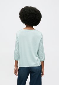 Άτομο με αφρο μαλλί που φορά ένα απαλό μπλε μακρυμάνικο ριπ πουκάμισο και σκούρες μπλε τζιν, στέκεται στραμμένο με την πλάτη προς το θεατή μπροστά σε ένα ουδέτερο φόντο.