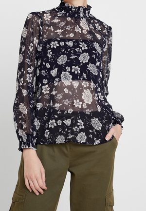 Blouse noire transparente à fleurs avec col montant volanté et poignets, portée avec un pantalon cargo vert olive.
