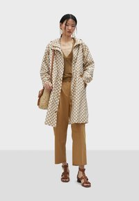 Cappotto beige a pois, top marrone e pantaloni a gamba larga coordinati. Sandali marroni con cinturini; porta una borsa di paglia marrone.