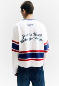 Fehér hosszú ujjú ing kék, piros és fehér vízszintes csíkokkal. A hátsó részén szöveg látható: "Jó az egészségnek, még jobb az egészségnek" és márkajelzés.