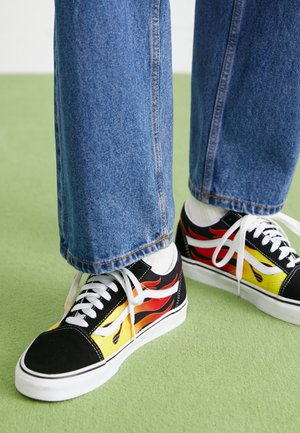 Czarne sneakersy z białymi sznurowadłami i podeszwami, ozdobione czerwono-żółtymi wzorami płomieni, noszone z niebieskimi dżinsami i białymi skarpetkami na zielonej podłodze.