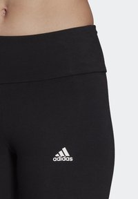 Svarta leggings i elastiskt tyg med ett brett midjeband. Har en liten vit Adidas-logotyp på nedre vänstra sidan.