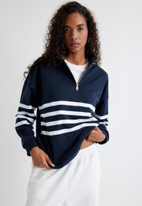 Sudadera navy blue con media cremallera y rayas horizontales blancas. Tela texturizada, corte relajado, mangas largas y detalle de cremallera plateada.