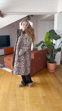 Casaco longo com padrão de leopardo e capuz, feito de tecido suave. Apresenta detalhes pretos e um emblema na manga. Usado com calças pretas e chinelos cinza.