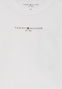 Fehér pamut póló kerek nyakkivágással. Jellemzője a "TOMMY HILFIGER" feliratos nyomtatott logó az "EST. 1985" alatt, valamint egy kis piros és kék zászló motívum.