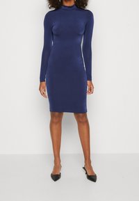 Tmavě modré šaty s dlouhým rukávem, stojáčkem, přiléhavým střihem a hladkou látkou. Nošeny s černými špičatými lodičkami.
