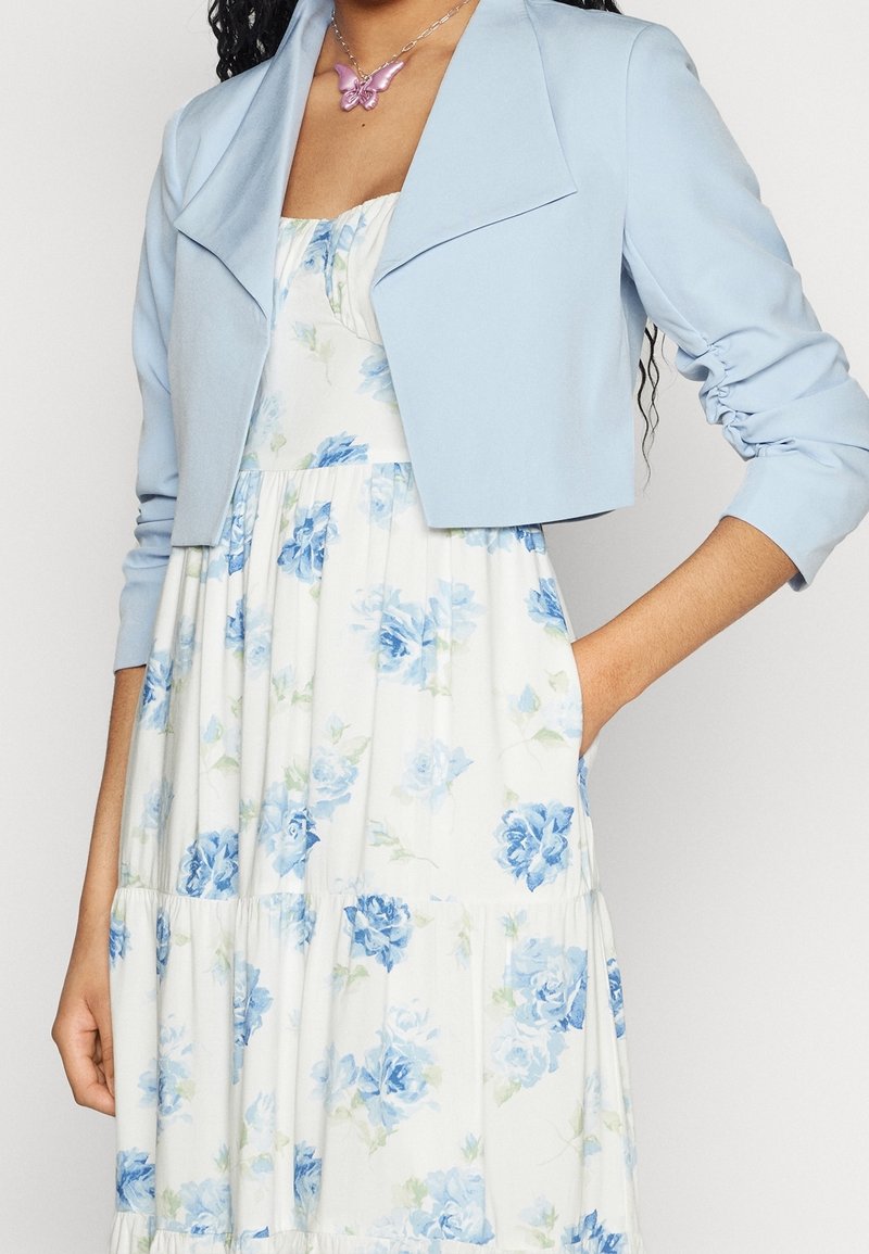 Veste courte bleu clair avec un col châle, tissu texturé et manches froncées, portée sur une robe blanche à motifs floraux et à volants.