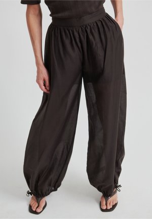 Mujer con pantalones negros sueltos y transparentes, con tobillos fruncidos y sandalias negras, de pie contra un fondo claro y liso.