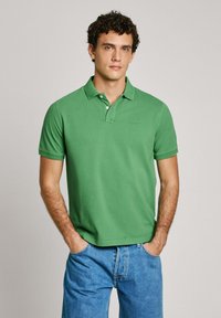 Green polo shirt with a collar, short sleeves, and a subtle logo on the chest. Worn with blue denim jeans, featuring a simple design.