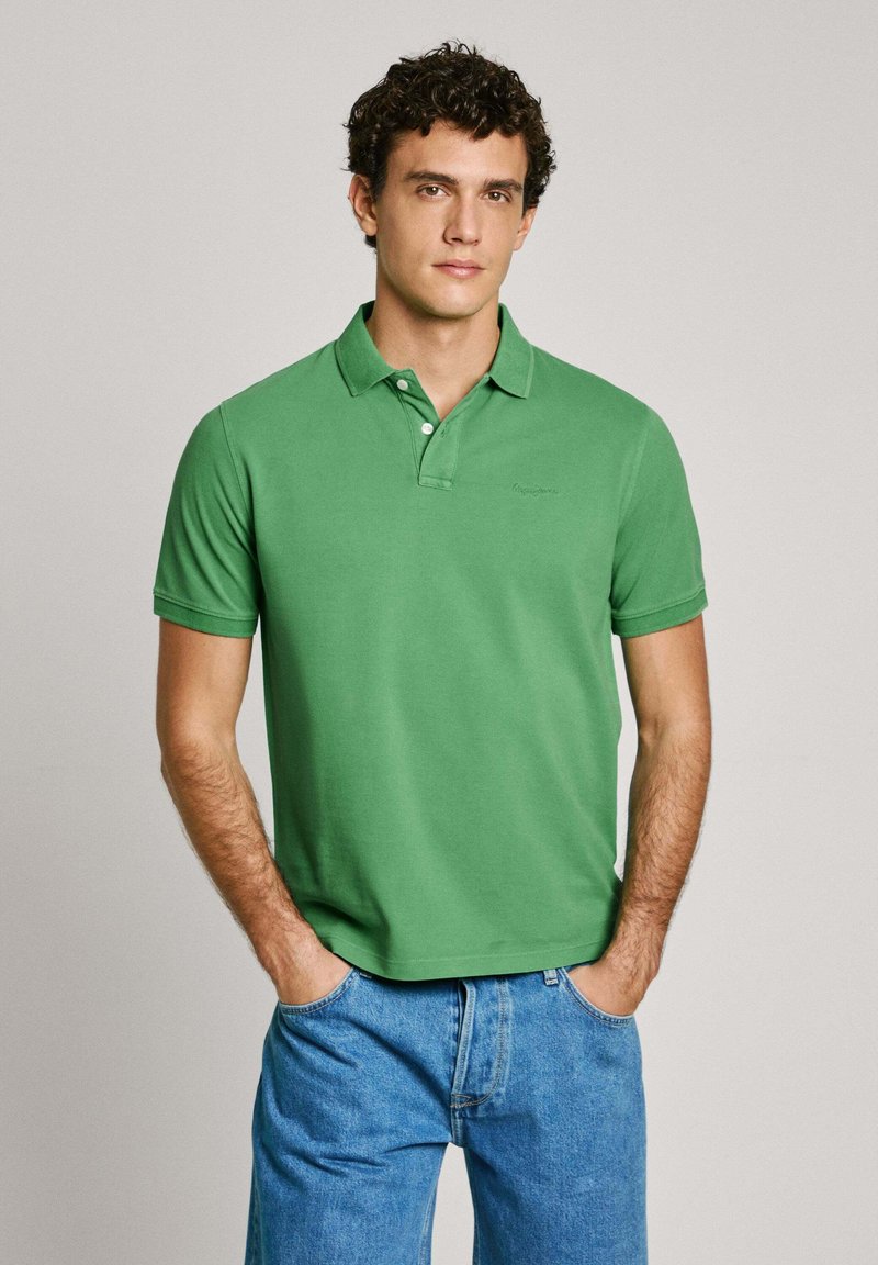 Green polo shirt with a collar, short sleeves, and a subtle logo on the chest. Worn with blue denim jeans, featuring a simple design.