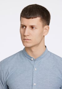 Camisa azul claro, de botones y con cuello mao. Tejido suave y texturizado con un sutil detalle de costura y botones blancos. Diseño ajustado.