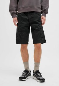 Shorts cargo noirs avec de multiples poches latérales, fabriqués en tissu léger. Portés avec des chaussettes beiges et des chaussures de sport noires.