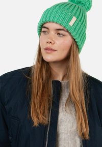 Green knitted beanie with a textured ribbed pattern and a pom-pom on top, featuring a small label. Worn with a dark jacket and light sweater.
