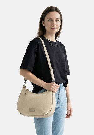 Sieviete, kas valkā melnu T-kreklu un gaiši zilas džinsas, turot bēša krāsas plecu somu ar regulējamu siksnu pāri ķermenim.