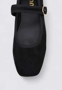 Zapato plano de ante negro con punta cuadrada, una correa negra estrecha en el empeine y un detalle de hebilla en tono dorado. Forro interior suave.