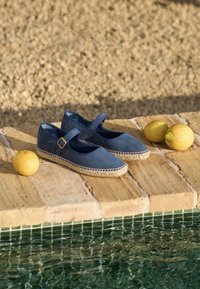 Blauwe suède Mary Jane schoenen met enkelbandje en espadrillezool, geplaatst op stenen tegels naast drie citroenen bij een zwembad.