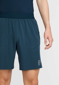 Mörkt blågröna sportshorts med elastisk midjeresår, utrustade med två sidofickor och en vit logotyp nära nederkanten. Mjukt, lätt tyg.