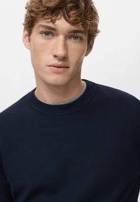 Marineblauer Strickpullover mit rundem Ausschnitt und gerippten Bündchen. Strukturierter Stoff mit dezentem Muster. Das Model hat kurze, lockige Haare.