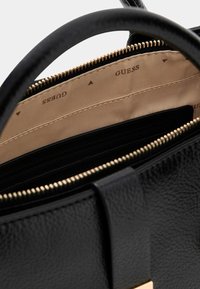 Czarna torebka o teksturowanej powierzchni z złotym zamkiem i beżową podszewką z logo marki "GUESS". Wnętrze zawiera liczne przegródki do organizacji.