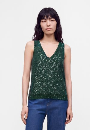 Donna con capelli neri lunghi indossa un top verde senza maniche con paillettes e jeans blu, in piedi davanti a uno sfondo chiaro e semplice.