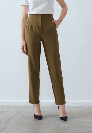 Frau trägt hochtaillierte, olivgrüne, zulaufende Hose und schwarze, spitze High Heels, steht vor einer schlichten hellen Wand, eine Hand in der Tasche.