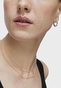 Collares de plata de ley con un diseño de cadena que se superponen alrededor del cuello. Pequeños pendientes de aro visibles en la oreja, sobre piel clara con pecas.