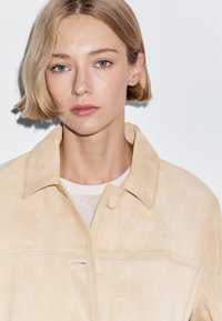 Giovane donna con capelli biondi corti e occhi verde chiaro che indossa una giacca in suede beige sopra un top bianco, espressione neutra, sfondo bianco uniforme.