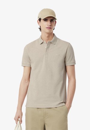 Beige pikétröja med korta ärmar och krage, med en liten broderad logotyp på bröstet. Tyget verkar mjukt och slätt.