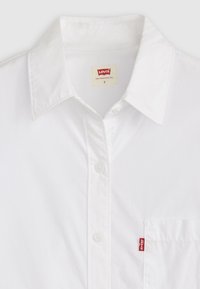 Chemise à col blanc en coton, dotée d'une patte de boutonnage sur le devant et d'une poche poitrine à gauche avec une étiquette de logo rouge.