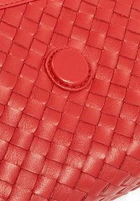 Czerwona skórzana torba o plecionej strukturze, z okrągłym zapięciem na guzik. Materiał wydaje się gładki i błyszczący, podkreślając złożone wzory tkane.