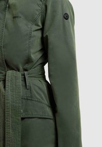 Oliviroheline pika varrukaga jakk kangasvöö, nööbitava klapiga tasku ja ümar must embleem ülemisel käel.