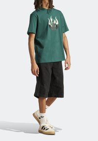 Maglietta verde in cotone con grafica a fiamma color crema e nero, abbinata a pantaloncini in denim neri e scarpe da ginnastica di colore chiaro con strisce.