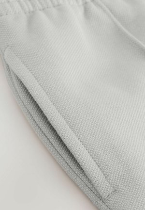 Tejido de color gris claro, texturizado y con una superficie suave. Presenta un ligero pliegue y una abertura lateral para un bolsillo, con una costura a lo largo del borde.