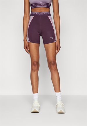 Atletul poartă pantaloni scurți sport Puma de culoare mov și lavandă, un top scurt asortat, cu șosete și adidași albi, stând în fața unui fundal alb.