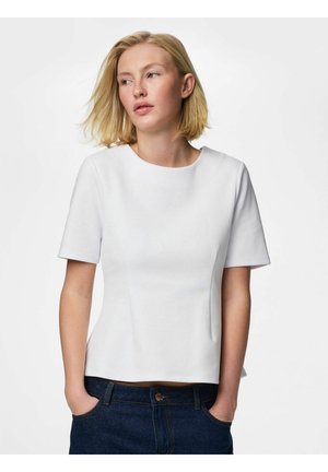 Donna bionda che indossa una maglietta bianca a maniche corte semplice e jeans blu scuro, con le mani nelle tasche, che guarda leggermente di lato.