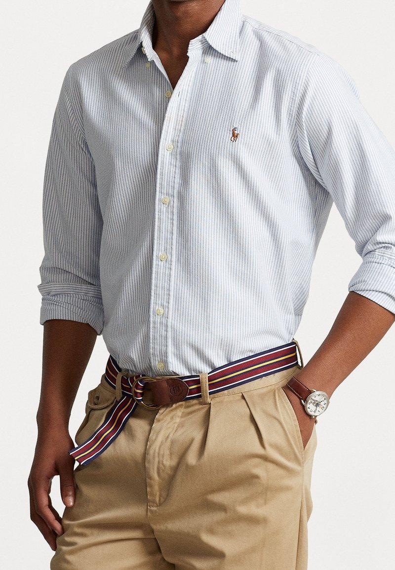 Blue and white striped button-down shirt, rolled sleeves, paired with beige trousers and a multicolored striped belt. Brown watch visible.