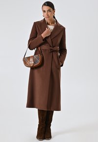 Cappotto in misto lana marrone con vita cinturata, rever oversize, tasche laterali, abbinato a una borsa con dettagli a quadri e stivali marroni con motivo.
