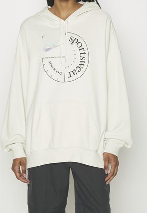 Osoba ubrana w za dużą, kremową bluzę z kapturem z czarną okrągłą grafiką „sportswear” i czarne spodnie, stojąca na jednolitym tle.