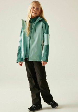 Junges Mädchen mit langen blonden Haaren, das eine türkis-grüne Jacke, schwarze Hose und schwarze Turnschuhe trägt, steht vor einem schlichten weißen Hintergrund.