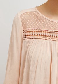 Persikofärgad blus med ett texturerat tyg, som har en spetspanel med cirkulära och prickiga mönster, långa ärmar och rund halsringning.