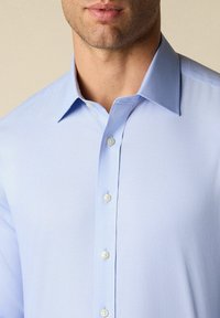 Lichtblauwe button-up shirt met een spread collar, zachte katoenen stof en vijf witte knopen langs de voorkant.