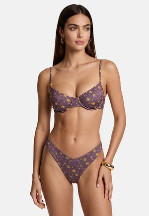 Donna che indossa un reggiseno a balconcino viola con motivo floreale e slip bikini a vita alta abbinati, con orecchini a cerchio dorati e braccialetto, sfondo neutro.