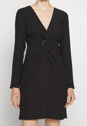 Kobieta ubrana w czarną sukienkę na długi rękaw z głębokim dekoltem w serek i srebrnym pierścieniem zbierającym tkaninę w talii.